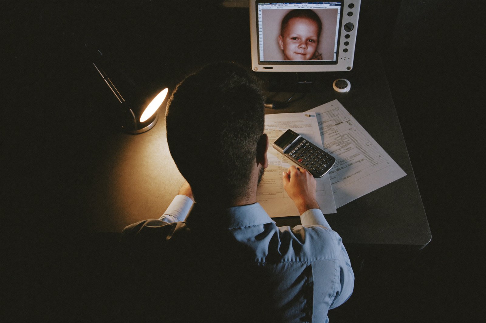 A man at a kitchen table late at night with printed stock charts and a calculator, baby monitor nearby