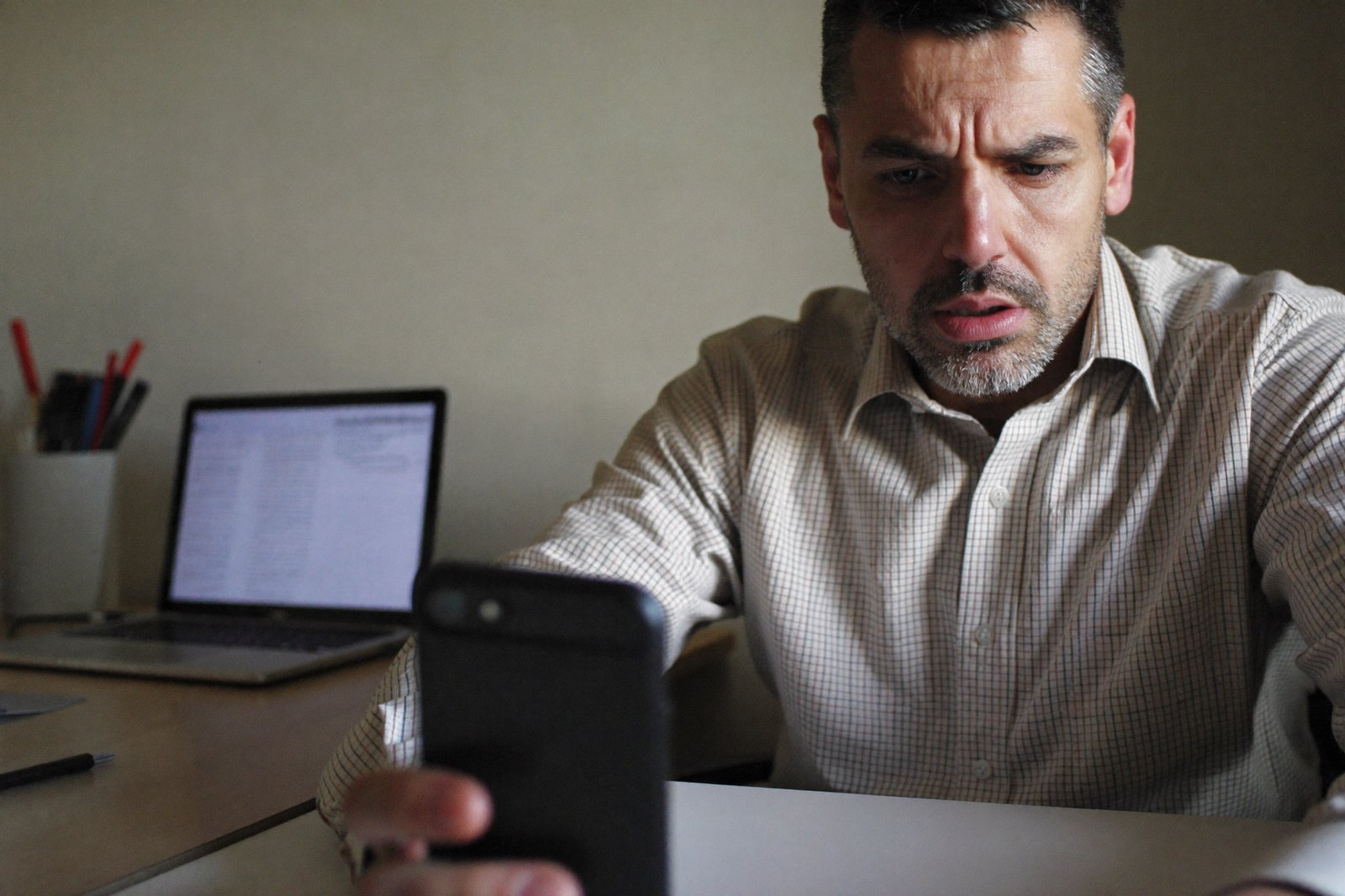 A man looking skeptically at his ringing phone at a desk, laptop open with an email inbox