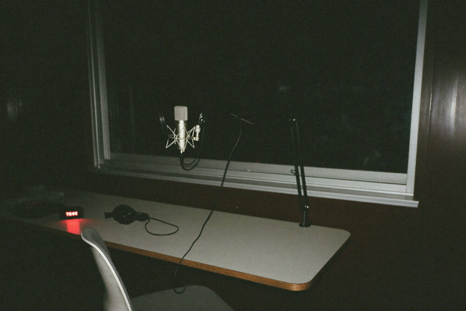 A podcast microphone and headphones on a desk in front of a dark window, recording light glowing red