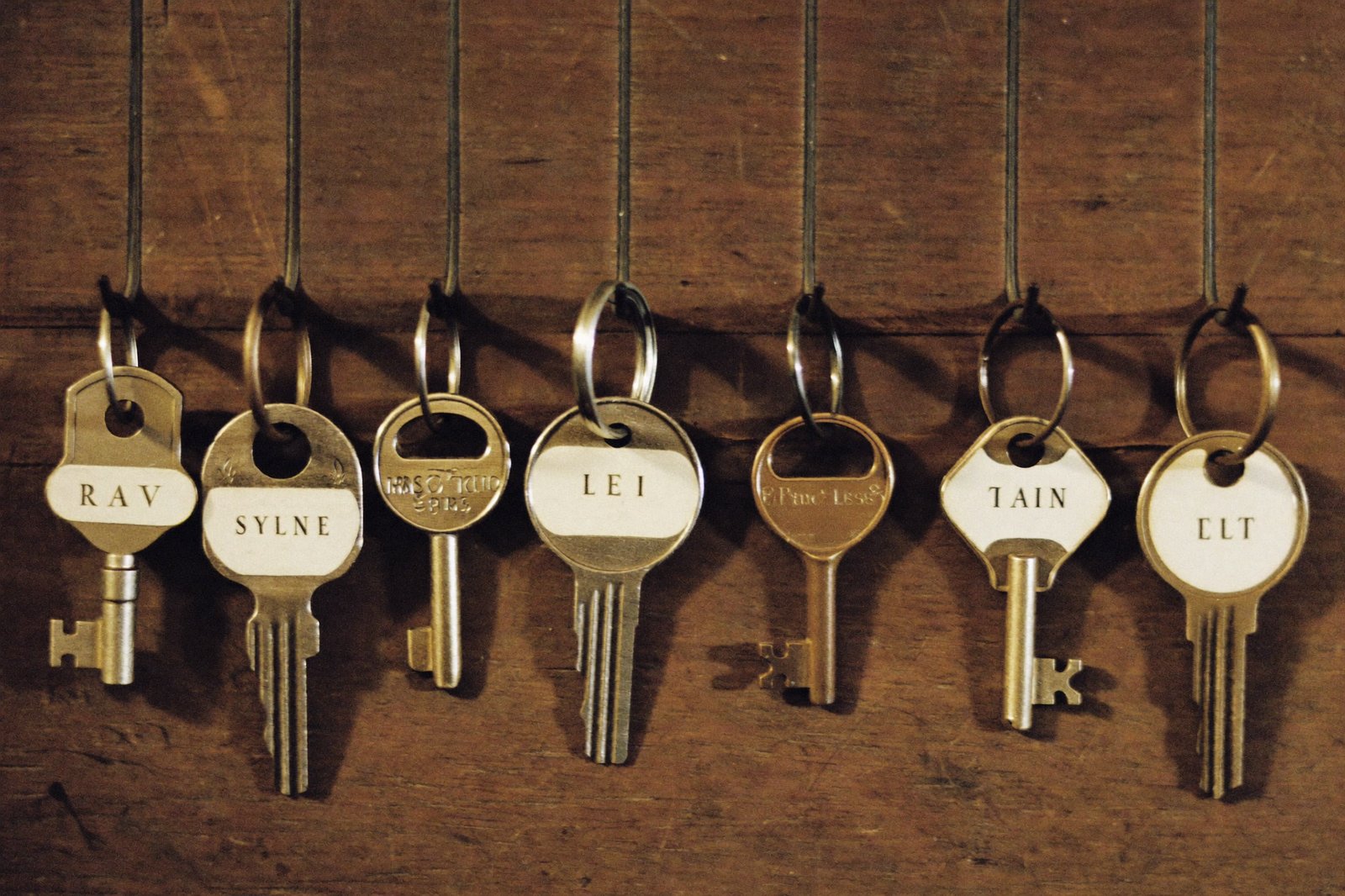 Seven labeled keys hanging in a row on a wooden board, one hook empty, warm side light