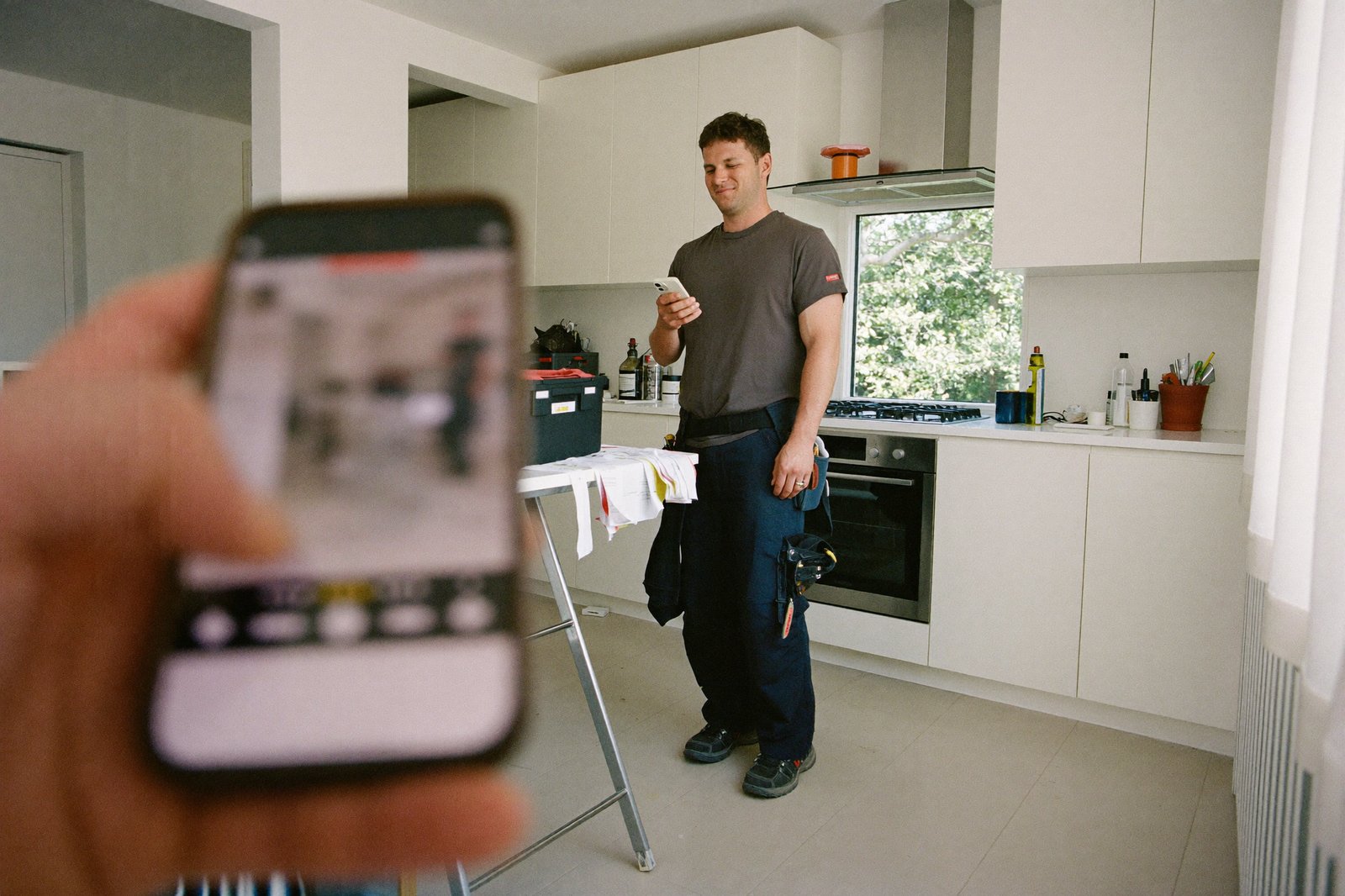 A tradesman in work clothes looking at his phone beside a toolbox in a bright kitchen, invoice papers on the counter
