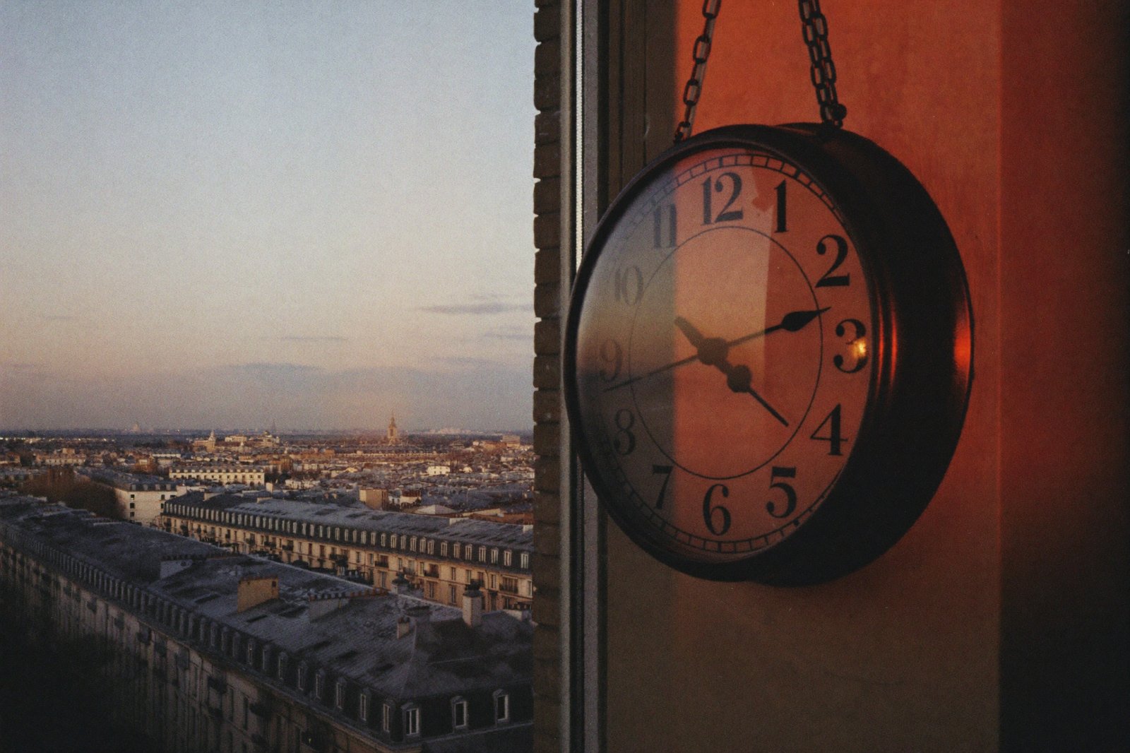 An old ticking wall clock beside a window at dusk, city rooftops fading into shadow