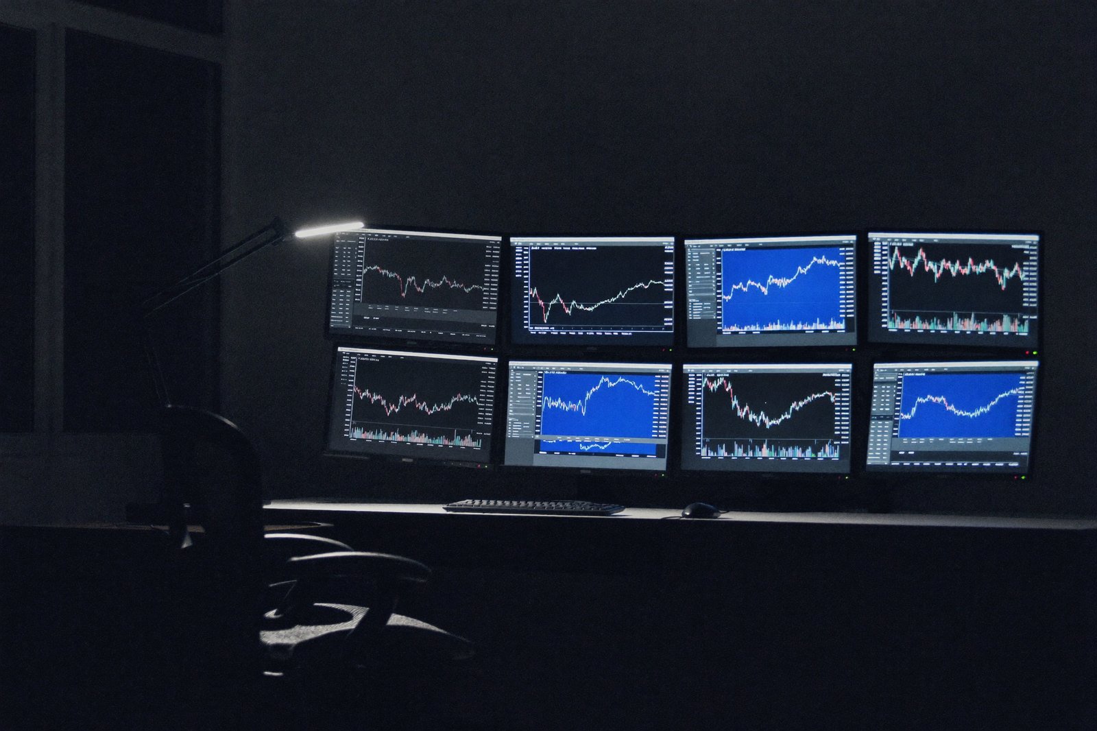 Multiple monitors of candlestick charts glowing in a dark home office, chair pushed back and empty